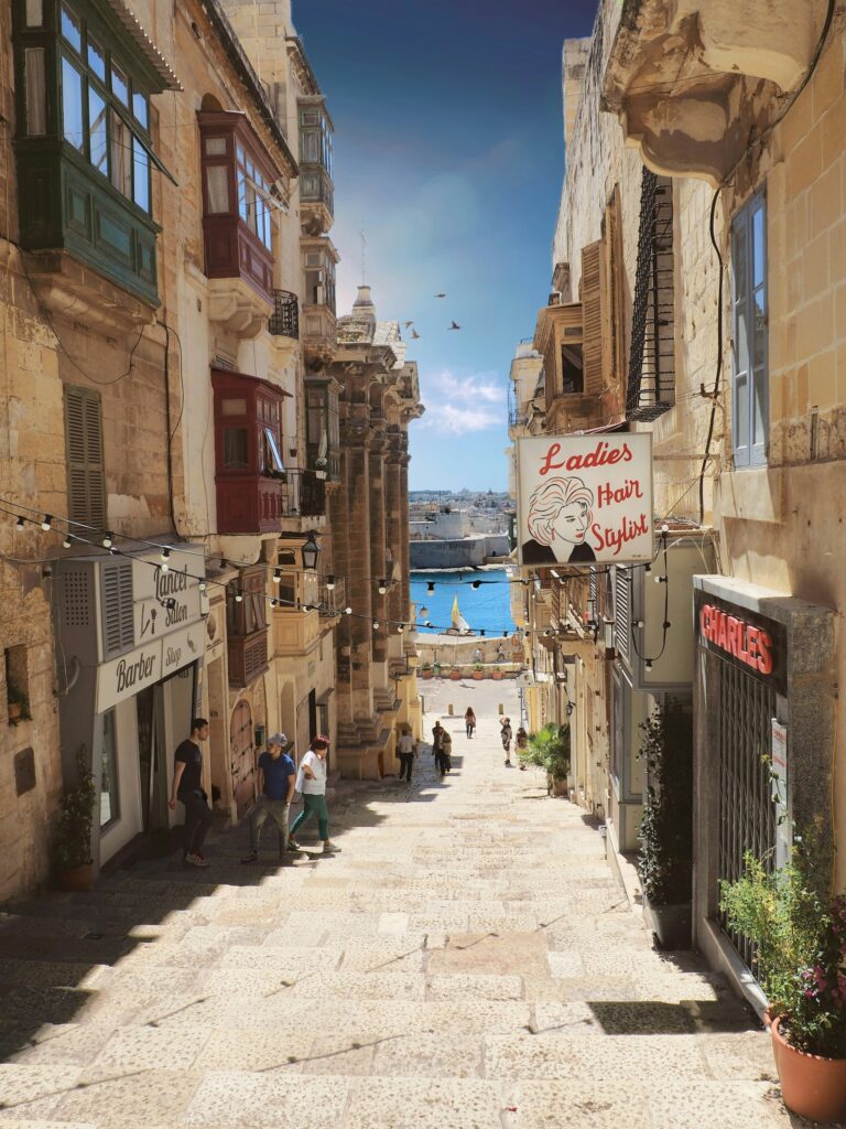 Słoneczna ulica w Valletcie z lokalnymi sklepami i przechodniami schodzącymi w kierunku Grand Harbour, Malta, październik